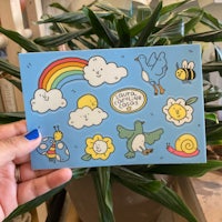 a hand holding a postcard with a rainbow, bees, and birds on it